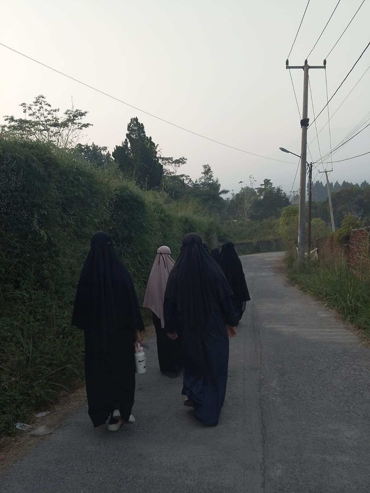 Dua santriwati berjalan berdampingan di jalan menuju ma'had pada pagi yang terang